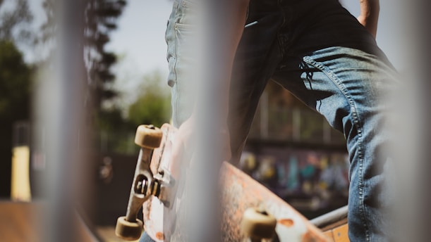 A close-up view featuring a skateboarder in jeans standing with one foot on a skateboard. The image is taken through a set of vertical bars, providing a sense of depth and framing. In the background, there's some graffiti art blurred out, suggesting an urban skate park setting.