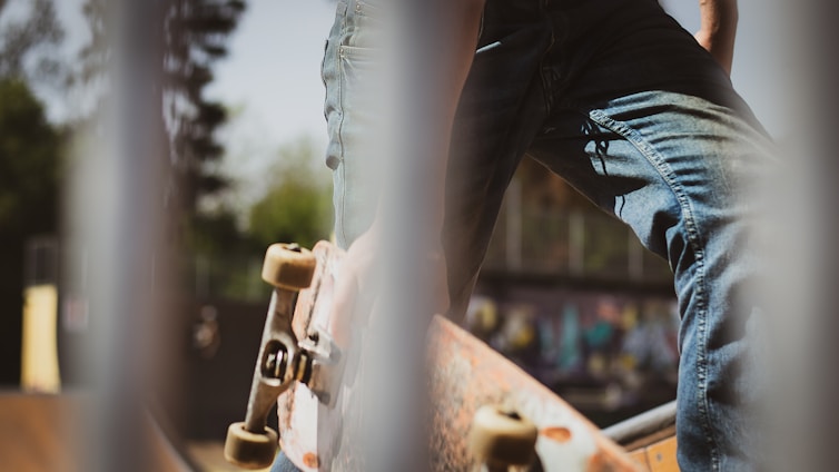 A close-up view featuring a skateboarder in jeans standing with one foot on a skateboard. The image is taken through a set of vertical bars, providing a sense of depth and framing. In the background, there's some graffiti art blurred out, suggesting an urban skate park setting.