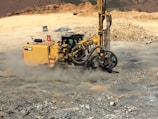 A large, yellow construction drill rig is operating on a rocky terrain, creating a cloud of dust. It is surrounded by loose stones and earth, with hills in the far background. The machine is equipped with hoses and drilling components, indicating its use in mining or construction work.