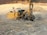 A large, yellow construction drill rig is operating on a rocky terrain, creating a cloud of dust. It is surrounded by loose stones and earth, with hills in the far background. The machine is equipped with hoses and drilling components, indicating its use in mining or construction work.