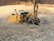 A large, yellow construction drill rig is operating on a rocky terrain, creating a cloud of dust. It is surrounded by loose stones and earth, with hills in the far background. The machine is equipped with hoses and drilling components, indicating its use in mining or construction work.