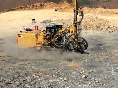 A large, yellow construction drill rig is operating on a rocky terrain, creating a cloud of dust. It is surrounded by loose stones and earth, with hills in the far background. The machine is equipped with hoses and drilling components, indicating its use in mining or construction work.