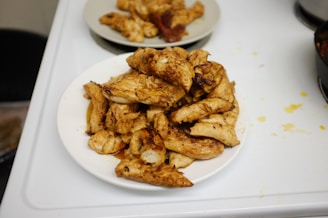 A white plate piled with grilled or roasted chicken pieces is placed on a kitchen countertop. The chicken appears to be seasoned and cooked, with a golden brown hue. In the background, another plate with additional chicken pieces is visible. The kitchen surface also shows some cooking splatters.