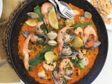 A large pan contains a vibrant paella dish, filled with rice, shrimp, mussels, clams, and lemon wedges. Fresh herbs are sprinkled on top, and there are green peppers and orange-colored rice adding a colorful touch. Two forks rest on the edge of the pan.