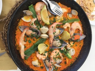 A large pan contains a vibrant paella dish, filled with rice, shrimp, mussels, clams, and lemon wedges. Fresh herbs are sprinkled on top, and there are green peppers and orange-colored rice adding a colorful touch. Two forks rest on the edge of the pan.