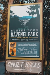 A wooden sign for Sunset Rock and Ravenel Park in Highlands, NC, is surrounded by foliage. The sign provides directions and information about the park, operated by the Highlands-Cashiers Land Trust, with an illustration of a scenic view including trees and mountains.