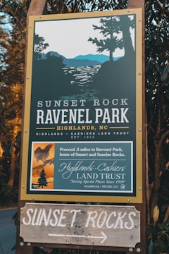 A wooden sign for Sunset Rock and Ravenel Park in Highlands, NC, is surrounded by foliage. The sign provides directions and information about the park, operated by the Highlands-Cashiers Land Trust, with an illustration of a scenic view including trees and mountains.