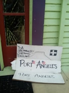 A cardboard sign is propped up against a colorful exterior wall, which is painted in pastel green and purple with a red-framed window. The sign has handwritten city names, including LA, San Francisco, Oakland, and Portland, with a larger font for 'Port Angeles' accompanied by small American flags.