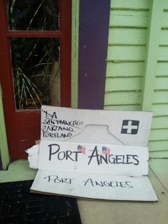 A cardboard sign is propped up against a colorful exterior wall, which is painted in pastel green and purple with a red-framed window. The sign has handwritten city names, including LA, San Francisco, Oakland, and Portland, with a larger font for 'Port Angeles' accompanied by small American flags.