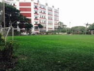 A large, well-maintained green field is in the foreground surrounded by a goalpost on the left. In the background, a multi-story building with red and white stripes stands tall with visible windows and air conditioning units. Several trees and plants surround the area, providing a lush, green landscape. A line of vehicles is parked near the building, and a few people are visible in the vicinity.