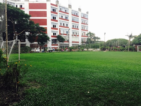 A large, well-maintained green field is in the foreground surrounded by a goalpost on the left. In the background, a multi-story building with red and white stripes stands tall with visible windows and air conditioning units. Several trees and plants surround the area, providing a lush, green landscape. A line of vehicles is parked near the building, and a few people are visible in the vicinity.