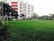 A large, well-maintained green field is in the foreground surrounded by a goalpost on the left. In the background, a multi-story building with red and white stripes stands tall with visible windows and air conditioning units. Several trees and plants surround the area, providing a lush, green landscape. A line of vehicles is parked near the building, and a few people are visible in the vicinity.
