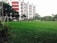 A large, well-maintained green field is in the foreground surrounded by a goalpost on the left. In the background, a multi-story building with red and white stripes stands tall with visible windows and air conditioning units. Several trees and plants surround the area, providing a lush, green landscape. A line of vehicles is parked near the building, and a few people are visible in the vicinity.