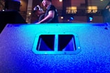 A large speaker dominates the foreground, its surface illuminated in vibrant blue light. In the background, two individuals are visible, one wearing headphones, standing on a stage with technical equipment around them. The lighting is dynamic, creating a lively atmosphere.