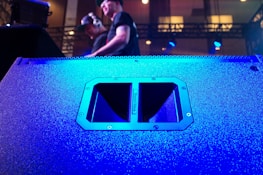 A large speaker dominates the foreground, its surface illuminated in vibrant blue light. In the background, two individuals are visible, one wearing headphones, standing on a stage with technical equipment around them. The lighting is dynamic, creating a lively atmosphere.