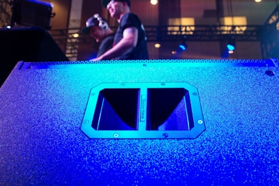 A large speaker dominates the foreground, its surface illuminated in vibrant blue light. In the background, two individuals are visible, one wearing headphones, standing on a stage with technical equipment around them. The lighting is dynamic, creating a lively atmosphere.