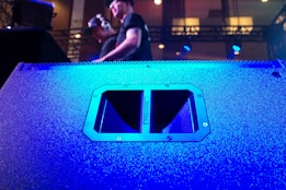 A large speaker dominates the foreground, its surface illuminated in vibrant blue light. In the background, two individuals are visible, one wearing headphones, standing on a stage with technical equipment around them. The lighting is dynamic, creating a lively atmosphere.