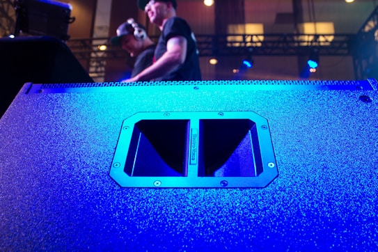 A large speaker dominates the foreground, its surface illuminated in vibrant blue light. In the background, two individuals are visible, one wearing headphones, standing on a stage with technical equipment around them. The lighting is dynamic, creating a lively atmosphere.