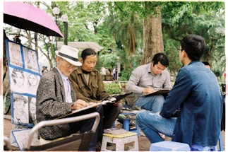 Several people are gathered in an outdoor setting, drawing or sketching on large pads of paper. One elderly man is wearing a hat and a patterned coat, focusing intently on his artwork. Other men are seated around him, also engaged in sketching. A pink umbrella is open above them, and nearby, portraits are displayed on a stand. The background is lush with green trees, creating a serene park atmosphere.