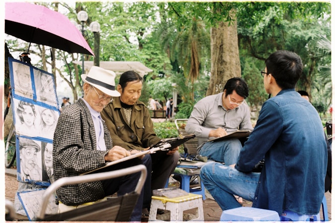 Several people are gathered in an outdoor setting, drawing or sketching on large pads of paper. One elderly man is wearing a hat and a patterned coat, focusing intently on his artwork. Other men are seated around him, also engaged in sketching. A pink umbrella is open above them, and nearby, portraits are displayed on a stand. The background is lush with green trees, creating a serene park atmosphere.