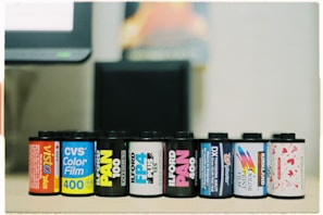 A row of eight film canisters are aligned on a surface. The canisters feature various colorful labels from different brands, including Agfa Vista, CVS Color Film, and Ilford Pan. The background appears slightly blurred with neutral tones, indicating a focus on the film canisters.