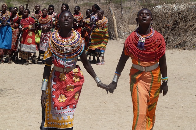 A group of people dressed in colorful traditional attire, with intricate beadwork and patterns, are gathered outdoors. Two women in the foreground are holding hands, wearing vibrant necklaces and robes with distinct cultural motifs. The background shows a group in similar attire, suggesting a community event.