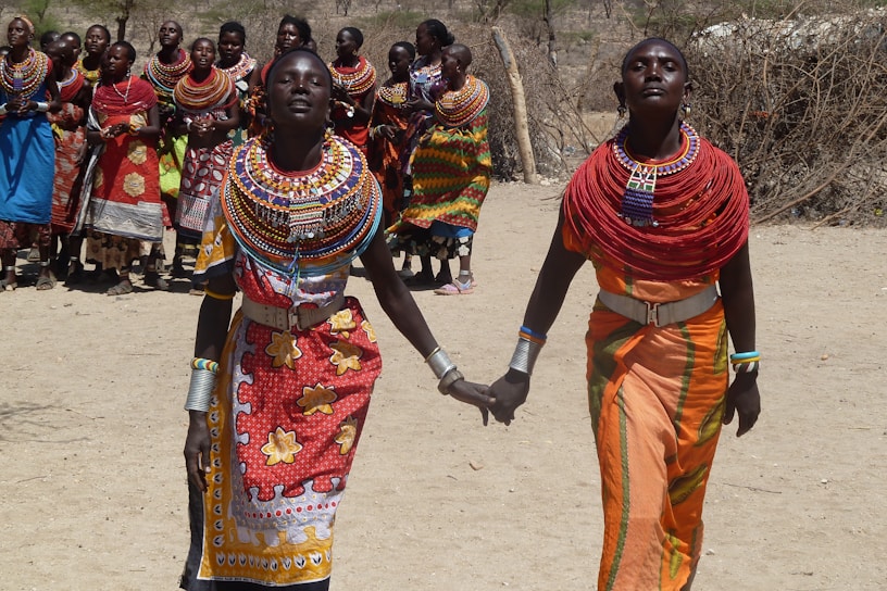 A group of people dressed in colorful traditional attire, with intricate beadwork and patterns, are gathered outdoors. Two women in the foreground are holding hands, wearing vibrant necklaces and robes with distinct cultural motifs. The background shows a group in similar attire, suggesting a community event.