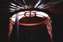 A view from inside a yurta showcasing an ornately decorated wooden frame and a partially open circular roof, allowing natural light to fill the interior. The intricate red and orange patterns on the wood add a cultural touch.