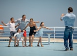 A group of five people, including two adults and three children, are posing playfully on what appears to be a ship deck. They are standing close together and smiling, with the ocean visible in the background. Another person is taking their photo.