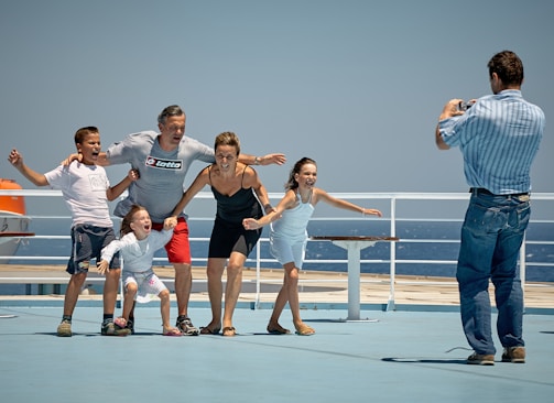 A group of five people, including two adults and three children, are posing playfully on what appears to be a ship deck. They are standing close together and smiling, with the ocean visible in the background. Another person is taking their photo.