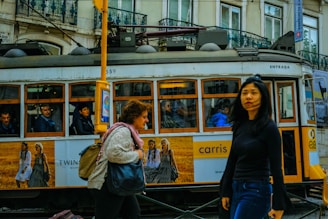 A vintage tram is moving through a city street, with people visible inside. Outside, two women walk by in opposite directions, one carrying a bag and the other gazing ahead. The tram features advertisements and is set against a backdrop of old buildings with intricate balconies.