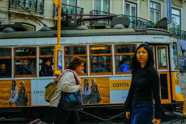 A vintage tram is moving through a city street, with people visible inside. Outside, two women walk by in opposite directions, one carrying a bag and the other gazing ahead. The tram features advertisements and is set against a backdrop of old buildings with intricate balconies.