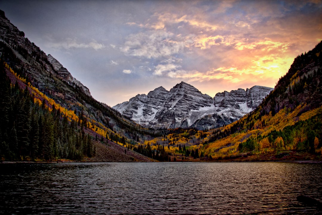 Photo by Mike Scheid of a majestic sunset behind craggy, rocky, snowy mountains. In the foreground is a calm lake, framed by evergreens and the golden aspen trees of the foothills surrounding the lake. 