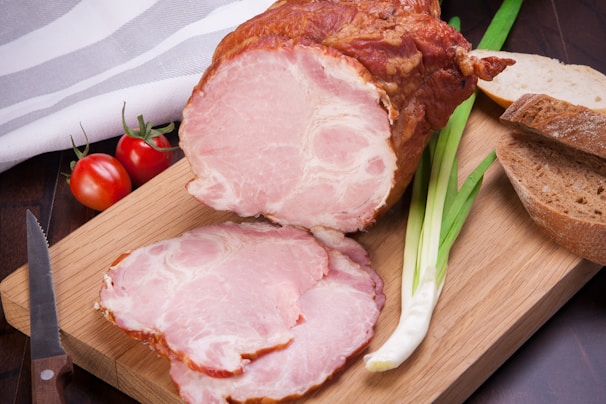 A wooden cutting board holds a large piece of smoked ham with a few slices cut from it. Adjacent to the ham are fresh vegetables including green onions and small red tomatoes. Slices of brown bread are also visible. A knife rests on the board, and a white-striped cloth is partially visible in the background.