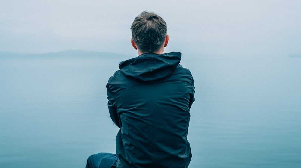 Homem com capuz preto sentado em frente ao oceano