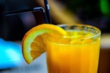 A refreshing glass of orange juice with ice cubes, garnished with a slice of orange on the rim, and two black straws positioned in the drink. The glass is set against a softly blurred background, hinting at a warm and relaxed atmosphere.