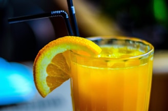 A refreshing glass of orange juice with ice cubes, garnished with a slice of orange on the rim, and two black straws positioned in the drink. The glass is set against a softly blurred background, hinting at a warm and relaxed atmosphere.