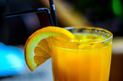 A refreshing glass of orange juice with ice cubes, garnished with a slice of orange on the rim, and two black straws positioned in the drink. The glass is set against a softly blurred background, hinting at a warm and relaxed atmosphere.