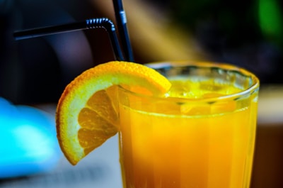 A refreshing glass of orange juice with ice cubes, garnished with a slice of orange on the rim, and two black straws positioned in the drink. The glass is set against a softly blurred background, hinting at a warm and relaxed atmosphere.