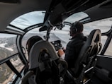 The cockpit of a helicopter with two people, a pilot and a passenger, flying over a scenic landscape with coastal views. The interior is sleek with glass windows providing a panoramic view. The pilot is wearing headphones and focused on controls.