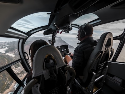 The cockpit of a helicopter with two people, a pilot and a passenger, flying over a scenic landscape with coastal views. The interior is sleek with glass windows providing a panoramic view. The pilot is wearing headphones and focused on controls.