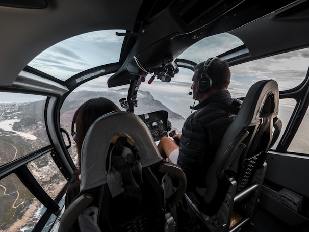 The cockpit of a helicopter with two people, a pilot and a passenger, flying over a scenic landscape with coastal views. The interior is sleek with glass windows providing a panoramic view. The pilot is wearing headphones and focused on controls.