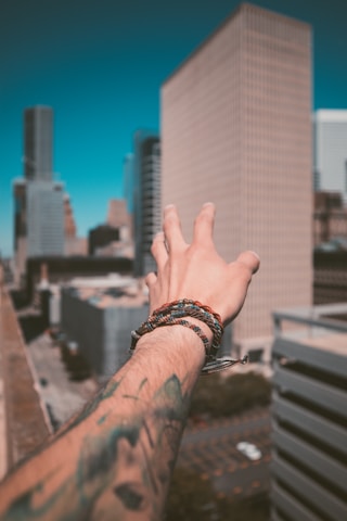 A tattooed arm reaching out towards a cityscape with tall buildings on a clear day. The wrist is adorned with multiple woven bracelets, and the perspective suggests the person is standing on a rooftop.