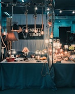 A dimly lit display booth with various vintage-style lamps and light bulbs, some hanging from ropes, others sitting on a table with a blue cloth. The setup has a retro and industrial vibe, with several decorative items and books placed around the lights. A sign reads 'MARTE LUMEN', suggesting a focus on lighting design.