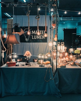 A dimly lit display booth with various vintage-style lamps and light bulbs, some hanging from ropes, others sitting on a table with a blue cloth. The setup has a retro and industrial vibe, with several decorative items and books placed around the lights. A sign reads 'MARTE LUMEN', suggesting a focus on lighting design.