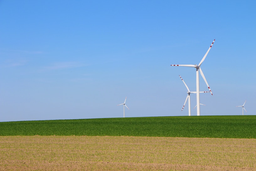 Wind turbines stand tall on a vast landscape, with a clear blue sky overhead. The foreground features patchy brown earth transitioning into lush green grass, creating a serene natural setting.