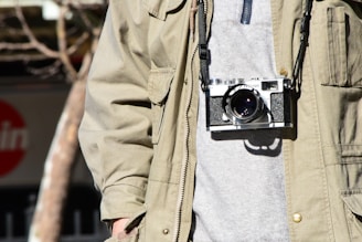 A person wearing a khaki jacket and grey top has a vintage film camera hanging from their neck. The camera is secured with a strap, and the scene has natural outdoor lighting with blurred background elements like trees.