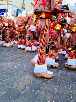 People in colorful and ornate costumes with detailed embroidery, including patterns of lions and other symbols, are gathered on a cobblestone street. Their outfits are vibrant with red, yellow, and black colors, complemented by decorative clogs and white knitted leg warmers.