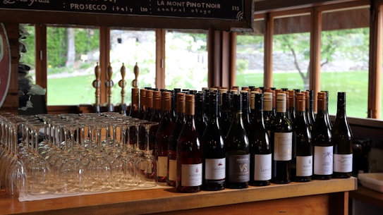 A collection of wine bottles arranged on a wooden counter, accompanied by neatly stacked wine glasses. The setting appears to be a cozy bar or tasting room, with large windows allowing natural light to illuminate the space and offer a view of a lush green outdoor area.