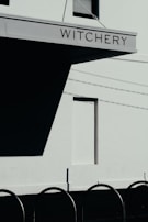 A modern building facade with a sign that reads 'Witchery' is prominently displayed. The structure casts a large shadow against a light-colored wall, beneath which lies a row of curved metal bars or bicycle racks. The image captures a clear day with visible shadows due to the sun.